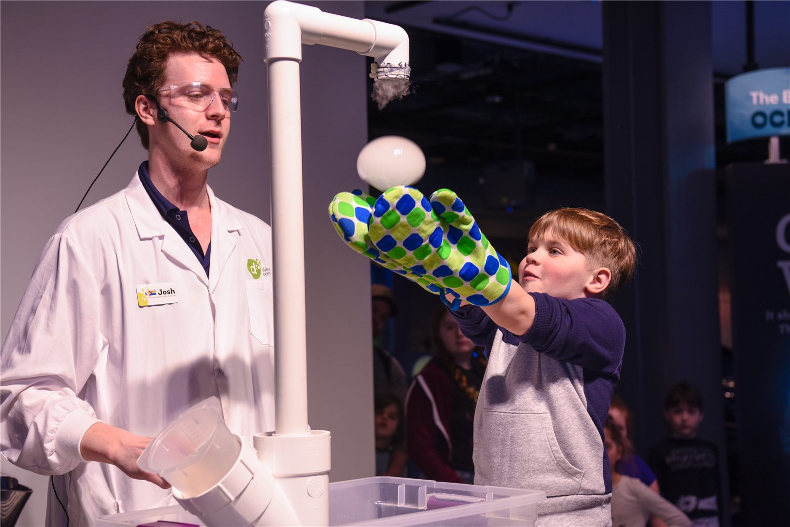 Educador do Discovery Centre conduz demonstração científica enquanto um menino com luvas coloridas segura uma bola flutuante